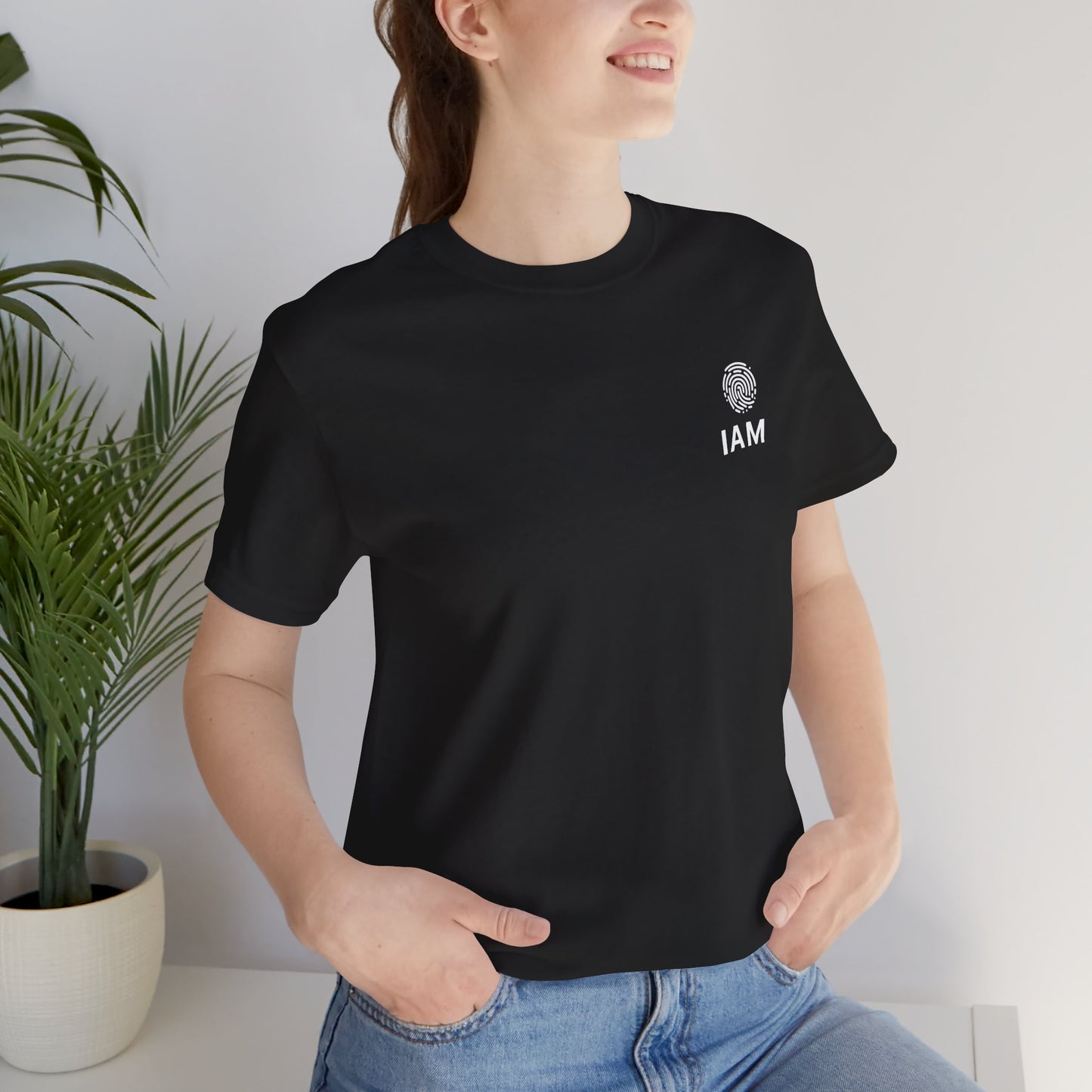 Person wearing a black t-shirt with 'IAM' logo and text, standing indoors with a plant in the background.