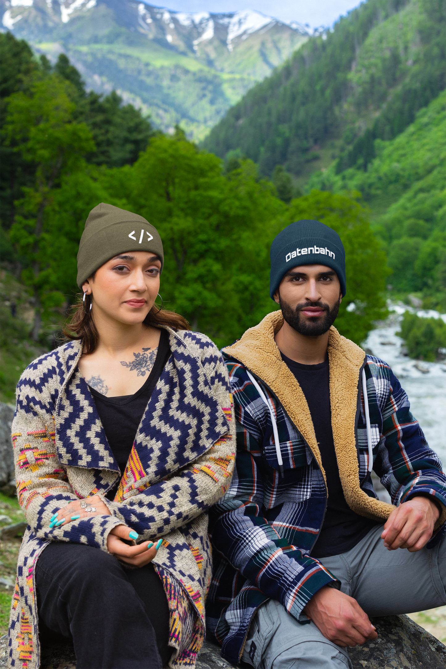 Two people sitting outdoors with mountains and a river in the background with beanies